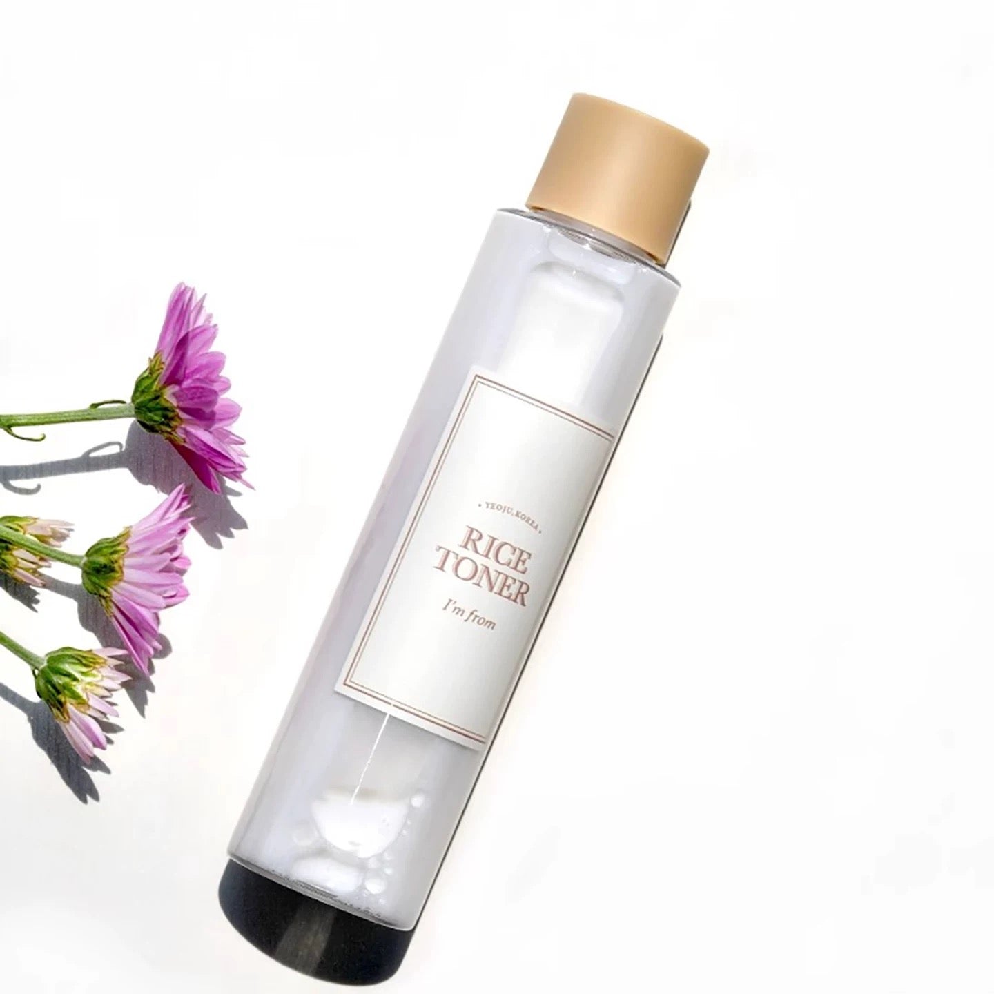 Bottle of rice toner with a beige cap and pink flowers on a white background